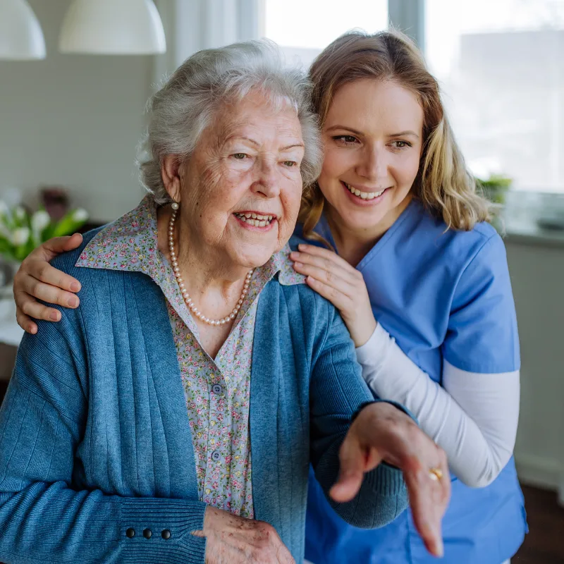 A smiling female caregiver in blue scrubs over a white long-sleeve shirt stands behind and gently rests her hands on the shoulders of an elderly woman wearing a pearl necklace and a blue cardigan over a floral top, as they both look off-camera with joyful expressions in a bright room.