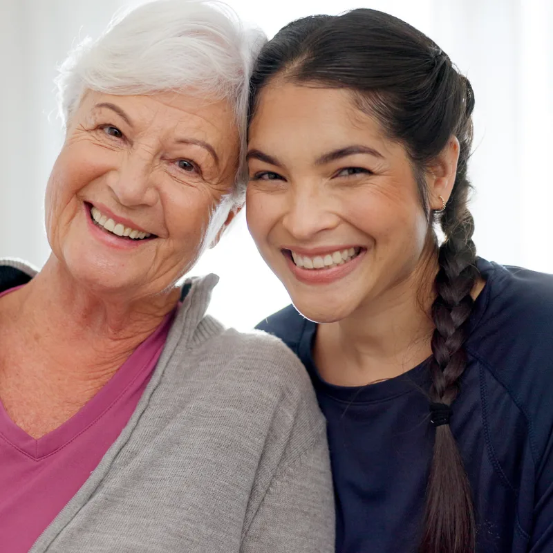 A close-up portrait of a smiling elderly woman with short white hair and a younger woman with a dark side-braid, leaning their heads together cheek-to-cheek and smiling broadly at the camera against a bright white background.