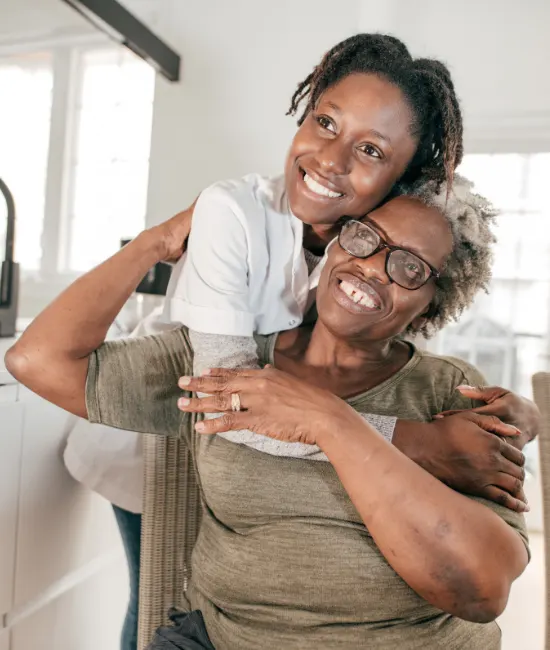 A smiling Black woman in a white medical top playfully wraps her arms around the shoulders of an older Black woman wearing glasses and an olive green shirt, both looking up and laughing joyfully in a bright home setting.