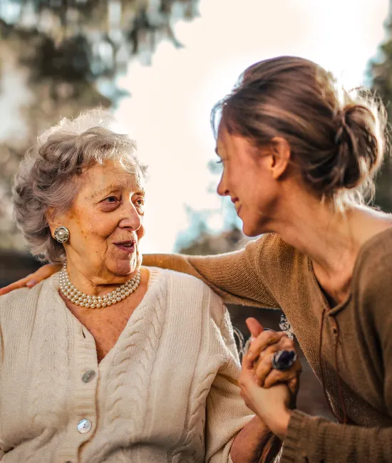 A younger woman in a brown knit sweater leans in to warmly embrace and hold the hand of an elegant elderly woman. The senior, wearing a pearl necklace and white cable-knit cardigan, smiles up at her with deep affection in a softly sunlit outdoor setting.