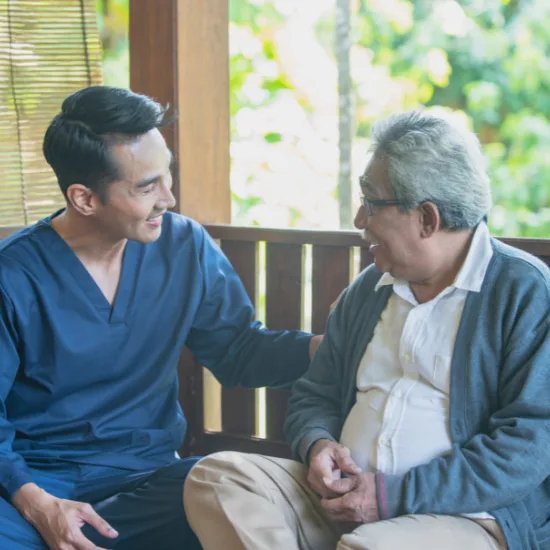 A smiling male caregiver in dark blue scrubs sits beside an elderly man with glasses on an outdoor wooden bench. The caregiver gently rests his hand on the senior's back as they share an uplifting, supportive conversation surrounded by lush green nature.