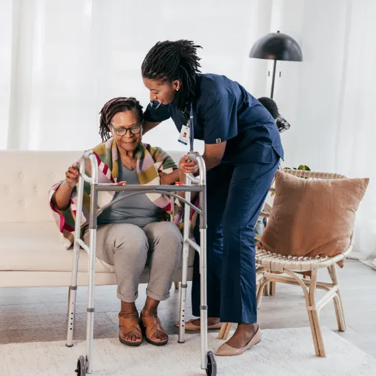 A careful nurse in navy blue scrubs bends down to guide an elderly woman's hands onto her silver walker, providing physical stability and hands-on support to help her stand up safely from a living room couch.