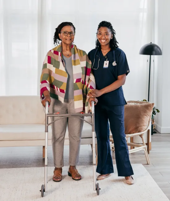A confident elderly woman stands upright with her walker, smiling alongside her supportive nurse in navy blue scrubs. The caregiver gently rests a hand on the walker, illustrating a successful journey toward regaining mobility and independence.
