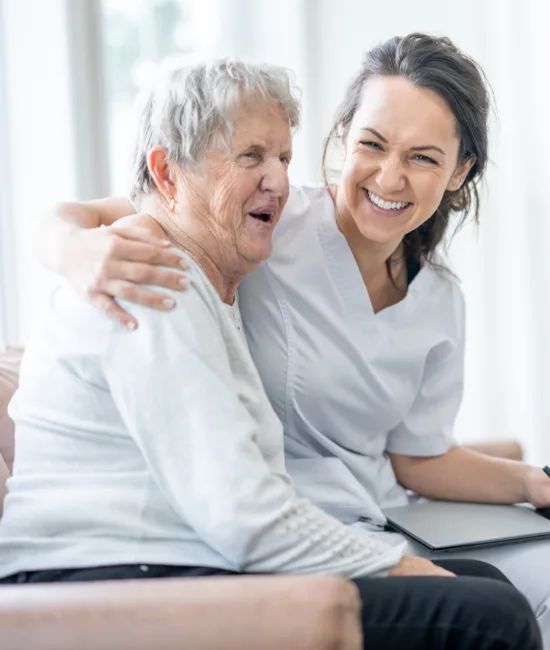 A cheerful nurse in a white scrub top sits on a couch with her arm around an elderly woman's shoulder. Both women are laughing heartily while looking at a laptop screen together in a bright, airy room.
