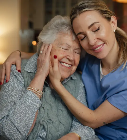 A gentle nurse in light blue scrubs cradles the face of an elderly woman in a tender, close embrace. Both women have their eyes closed in a moment of pure contentment and emotional connection.