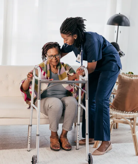 A Black female nurse in dark blue scrubs leans down to assist an elderly woman with glasses and a colorful plaid blanket as she attempts to stand up from a white sofa using a silver walker.